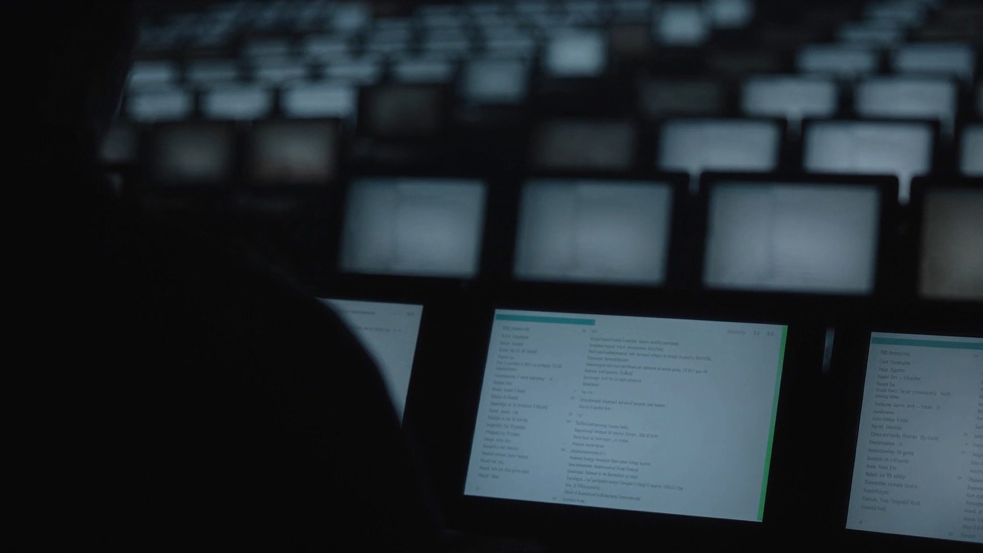A silhouetted figure in a dark room facing rows of glowing screens filled with text threads
